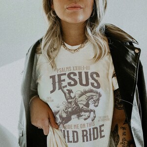 K&ouml;nnte beinhalten: Wei&szlig;es T-Shirt mit einer braunen Grafik eines Cowboys, der ein Pferd reitet. Der Text "PSALMS XXIII:I-III JESUS GUIDE ME ON THIS WILD RIDE" ist auf das T-Shirt gedruckt.