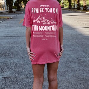 May include: A watermelon-colored t-shirt with the text "SO I WILL PRAISE YOU ON THE MOUNTAIN" in white. The design includes a mountain and trees graphic. The text "PSALM 121" is also visible.
