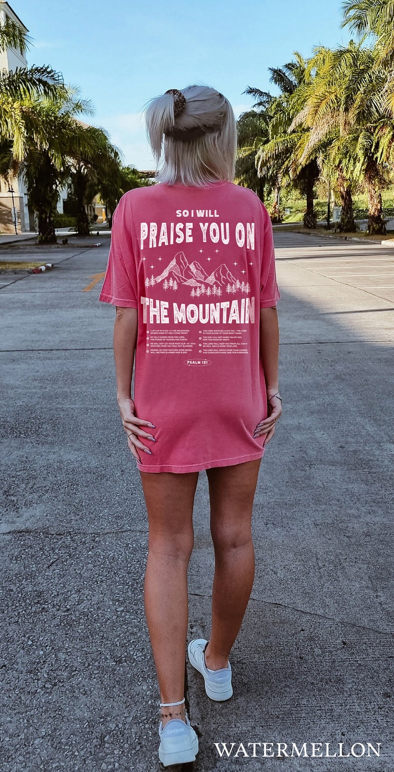 May include: A watermelon-colored t-shirt with the text "SO I WILL PRAISE YOU ON THE MOUNTAIN" in white. The design includes a mountain and trees graphic. The text "PSALM 121" is also visible.
