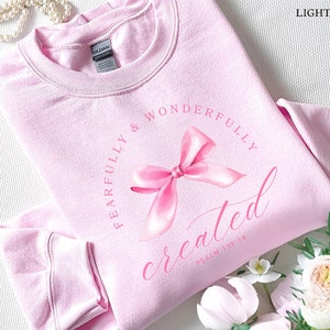 May include: Light pink sweatshirt with a pink bow and the text "Fearfully & Wonderfully Created" with the scripture reference "Psalm 139:14".