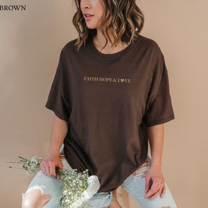 May include: A brown oversized t-shirt with the text "FAITH HOPE & LOVE" printed in gold on the front. The shirt is being worn by a person with long brown hair.