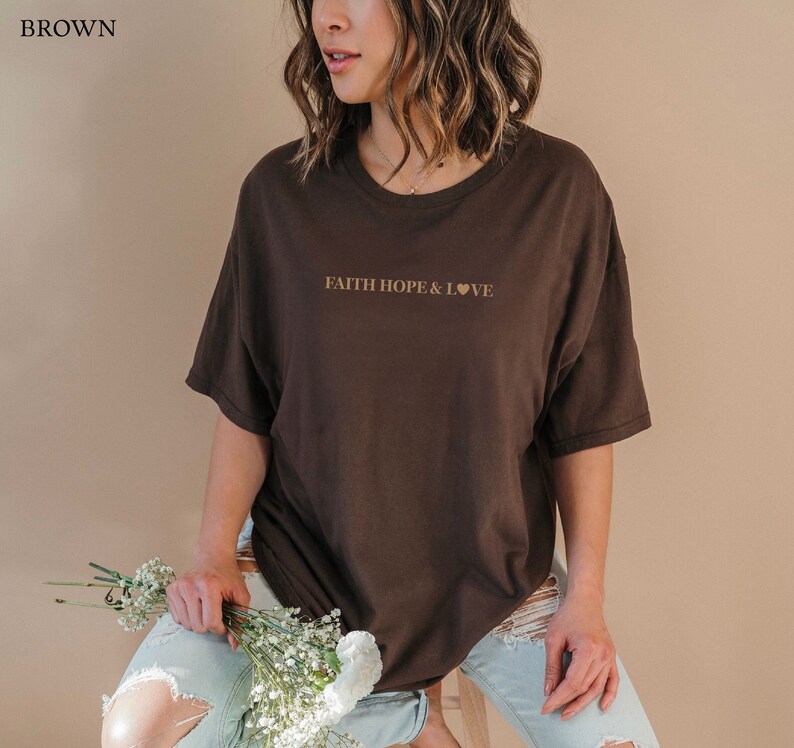 May include: A brown oversized t-shirt with the text "FAITH HOPE & LOVE" printed in gold on the front. The shirt is being worn by a person with long brown hair.