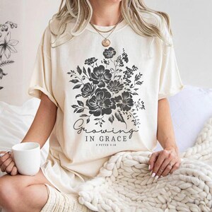 May include: A woman wearing a cream colored t-shirt with a black floral design and the text "Growing in Grace 2 Peter 3:18".