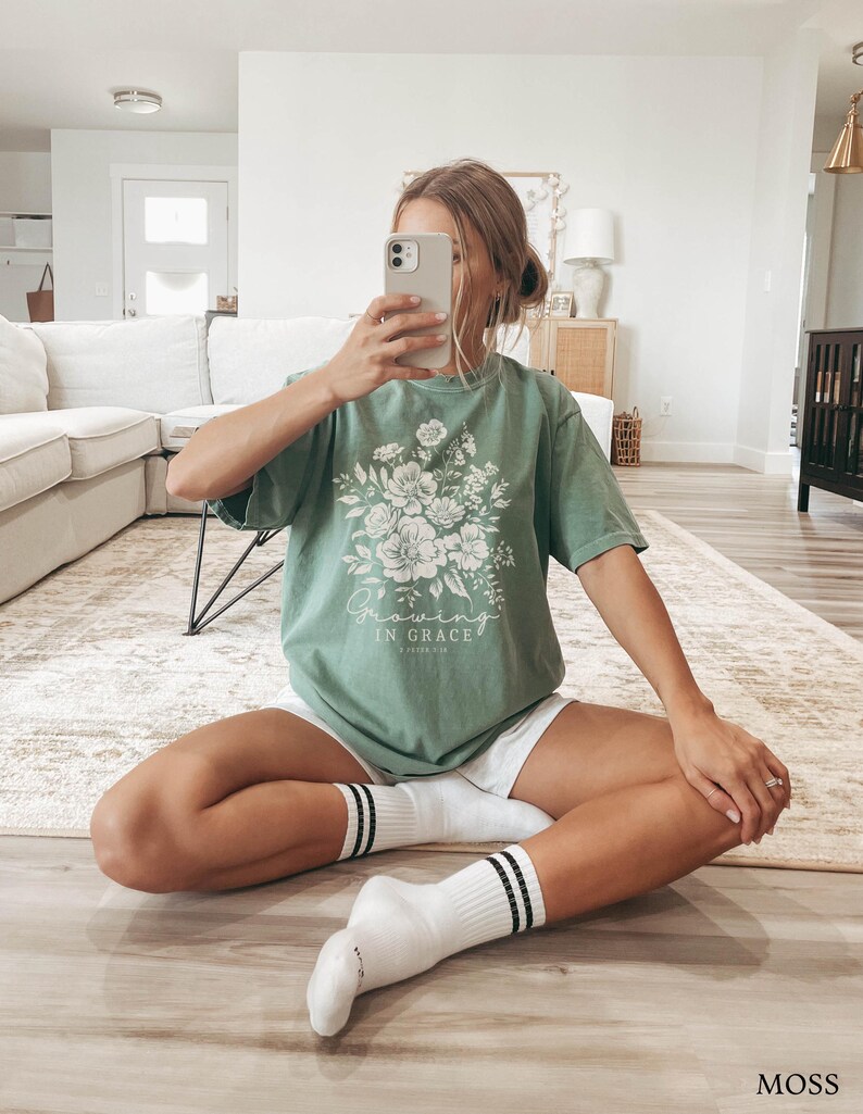 May include: A woman wearing a green t-shirt with a floral design and the text "Growing in Grace" printed on it. She is wearing white shorts and white socks with black stripes.