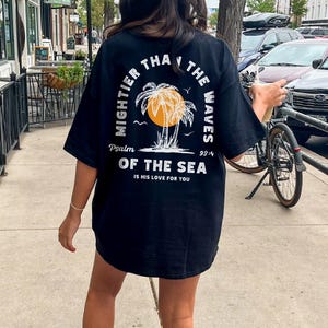 Puede incluir: Camiseta negra con un gr&aacute;fico blanco de palmeras y el texto "Mightier than the waves Psalm 93:4 Of the sea is his love for you".