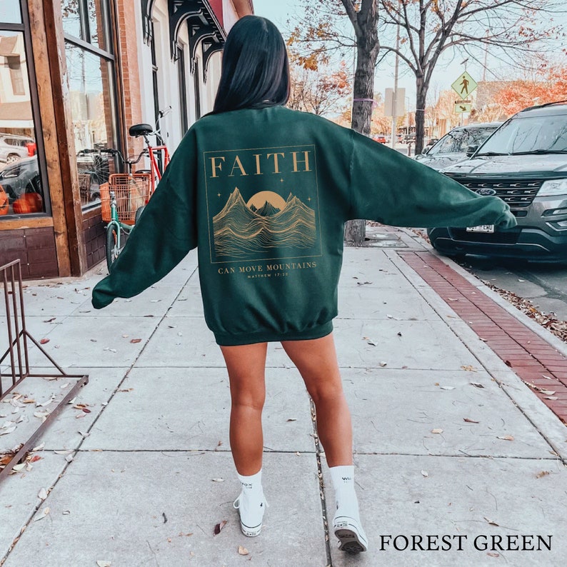 May include: Forest green sweatshirt with a gold graphic of mountains and a sun. The graphic includes the text "FAITH CAN MOVE MOUNTAINS MATTHEW 17:20".