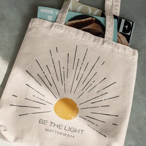 Op de afbeelding: Cr&egrave;mekleurige draagtas met een zwart zonnestraalontwerp en een gele zon. De tekst "BE THE LIGHT" en "MATTHEW 5:14" zijn eronder gedrukt. De tas bevat tijdschriften.