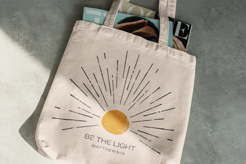Op de afbeelding: Cr&egrave;mekleurige draagtas met een zwart zonnestraalontwerp en een gele zon. De tekst "BE THE LIGHT" en "MATTHEW 5:14" zijn eronder gedrukt. De tas bevat tijdschriften.