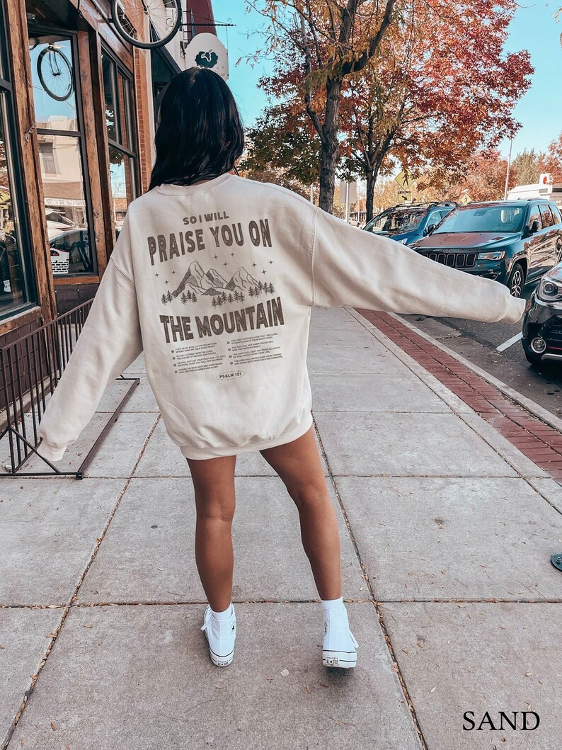 May include: A beige sweatshirt with a graphic print of mountains and the text "So I will praise you on the mountain".