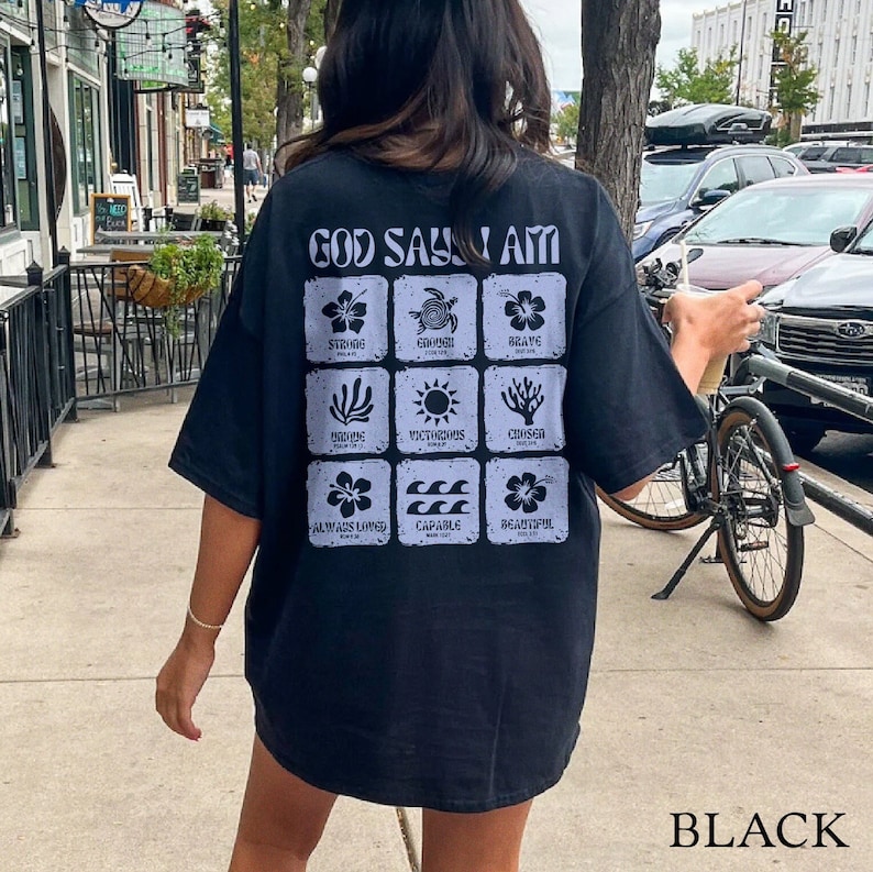 May include: Black t-shirt with a graphic print that says "God Says I Am" and features a grid of nine squares with different images and words in each square. The words include "Strong", "Enough", "Brave", "Chosen", "Victorious", "Capable", "Always Loved", and "Beautiful".
