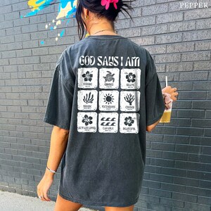 May include: A black t-shirt with a white graphic print that says "God Says I Am" with a grid of nine squares. Each square has a different image and word, including "Strong", "Enough", "Brave", "Chosen", "Victorious", "Always Loved", "Capable", and "Beautiful".