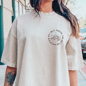 May include: Oversized beige t-shirt with a circular graphic on the upper back. The graphic features a mountain range, trees, stars, and the text "SO I WILL PRAISE YOU ON THE MOUNTAIN". The shirt has short sleeves and a crew neck.