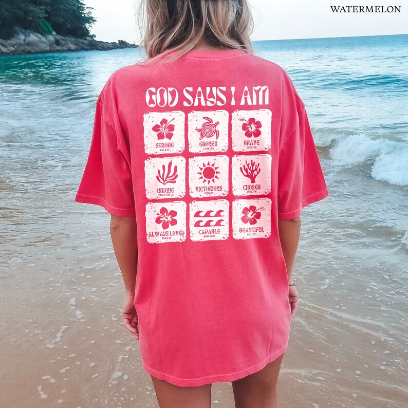 May include: A pink t-shirt with a white graphic that says "God Says I Am" and features a grid of nine squares with different images and words, including "Strong", "Brave", "Victorious", "Chosen", "Always Loved", and "Capable".