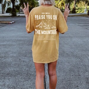 May include: Mustard-colored t-shirt with the text "SO I WILL PRAISE YOU ON THE MOUNTAIN" and a mountain graphic. The person is wearing a black baseball cap with "CA" on it and white sneakers.