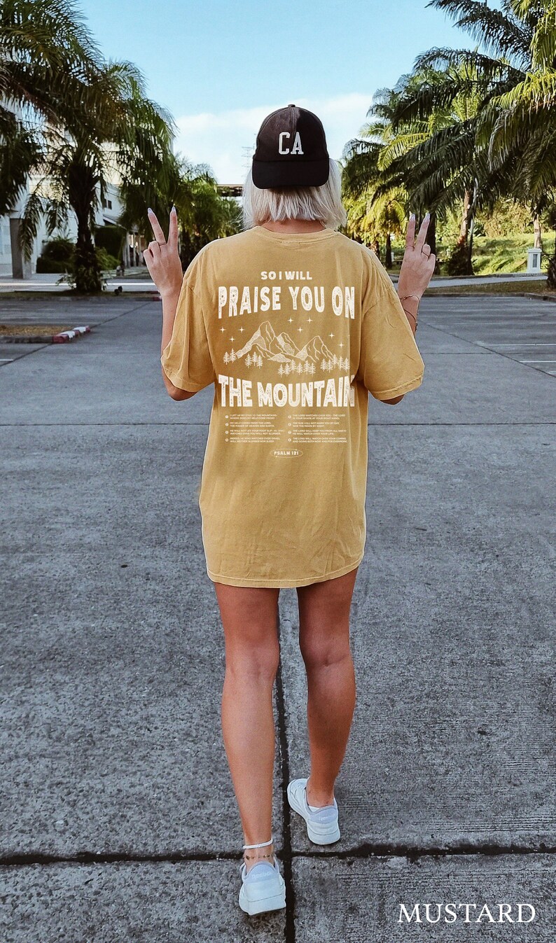 May include: Mustard-colored t-shirt with the text "SO I WILL PRAISE YOU ON THE MOUNTAIN" and a mountain graphic. The person is wearing a black baseball cap with "CA" on it and white sneakers.