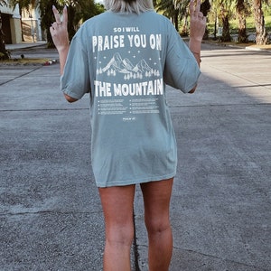 May include: A light green t-shirt with the text "SO I WILL PRAISE YOU ON THE MOUNTAIN" and a mountain graphic. The person is wearing a black baseball cap with "CA" on it and white sneakers.