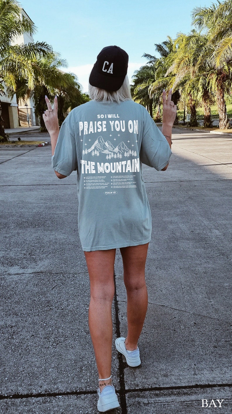 May include: A light green t-shirt with the text "SO I WILL PRAISE YOU ON THE MOUNTAIN" and a mountain graphic. The person is wearing a black baseball cap with "CA" on it and white sneakers.