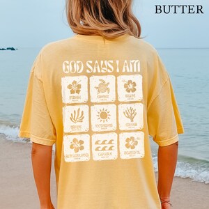 May include: A yellow t-shirt with the text "God Says I Am" printed on the back. The text is surrounded by a grid of nine squares, each containing a different image and word, such as "Strong", "Enough", "Brave", "Unique", "Victorious", "Chosen", "Always Loved", "Capable", and "Beautiful".