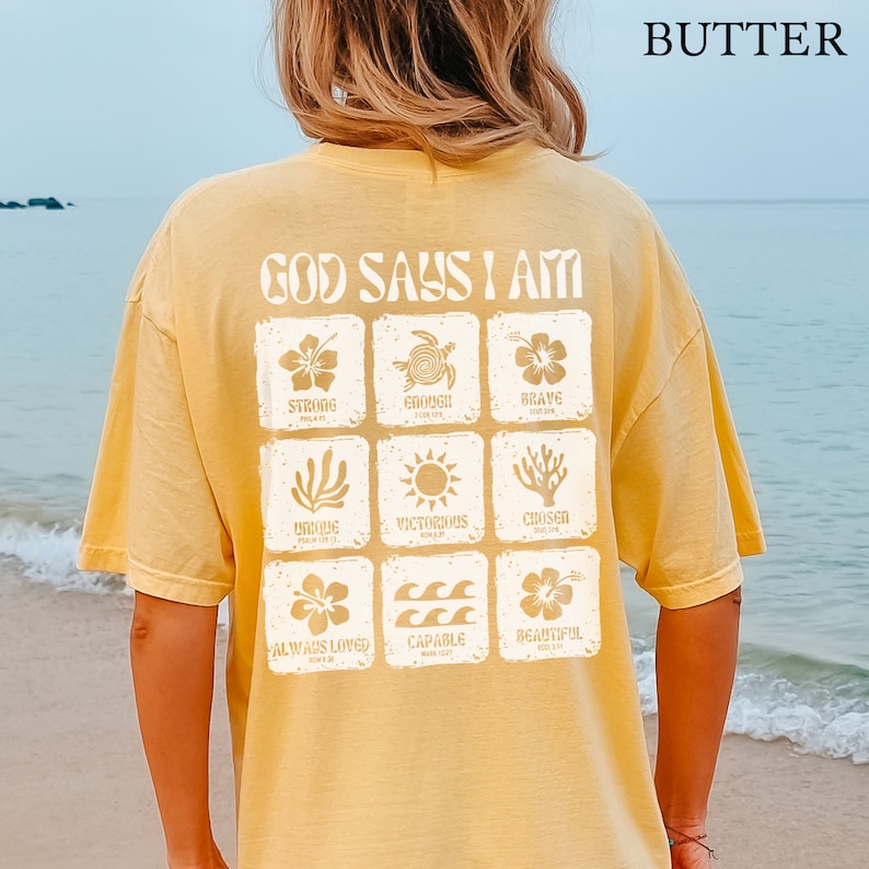 May include: A yellow t-shirt with the text "God Says I Am" printed on the back. The text is surrounded by a grid of nine squares, each containing a different image and word, such as "Strong", "Enough", "Brave", "Unique", "Victorious", "Chosen", "Always Loved", "Capable", and "Beautiful".