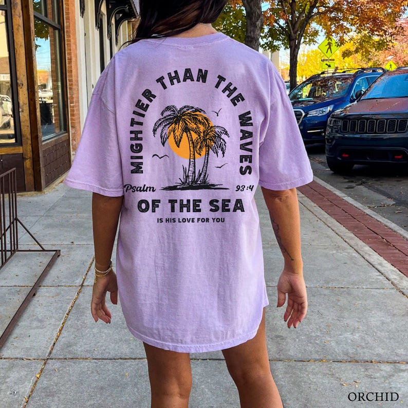 Puede incluir: Una camiseta lavanda con un estampado gr&aacute;fico de palmeras y un sol. El texto en la camiseta dice "Mightier Than The Waves Psalm 93:4 Of The Sea Is His Love For You".