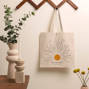 Op de afbeelding: Beige draagtas met een zonnestralenontwerp en de tekst "BE THE LIGHT" in zwart. De tas hangt aan een houten wandrek. Een roze vaas met eucalyptus en een kaars staan op een houten tafel.