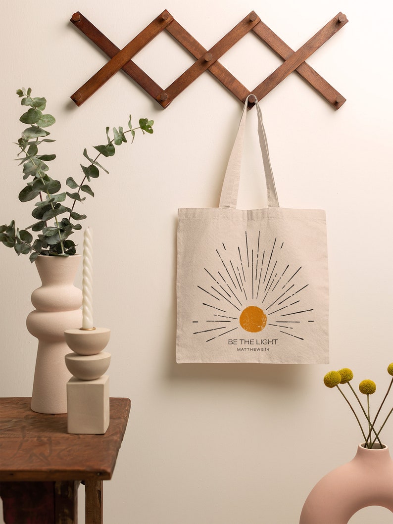 Op de afbeelding: Beige draagtas met een zonnestralenontwerp en de tekst "BE THE LIGHT" in zwart. De tas hangt aan een houten wandrek. Een roze vaas met eucalyptus en een kaars staan op een houten tafel.