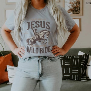 K&ouml;nnte beinhalten: Ein hellblaues T-Shirt mit einem Grafikdruck eines Cowboys, der ein Pferd reitet, mit dem Text "Psalms XXIII:4", "Jesus", "Ride On My" und "Wild Ride".