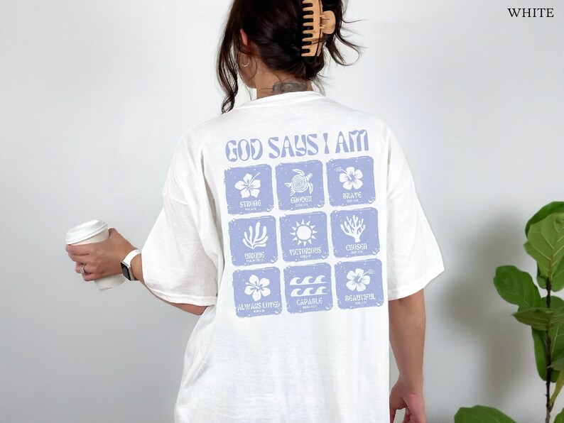 May include: White t-shirt with a blue graphic print that says "God Says I Am" and includes nine squares with different images and words, including "Strong", "Composed", "Brave", "Loving", "Victorious", "Courage", "Always Loved", "Capable", and "Beautiful".