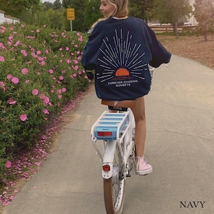 May include: A person riding a white electric bicycle with a blue basket. The person is wearing a navy blue sweatshirt with a graphic of a sunset and the text "FOREVER CHASING SUNSETS". The sweatshirt is navy blue.