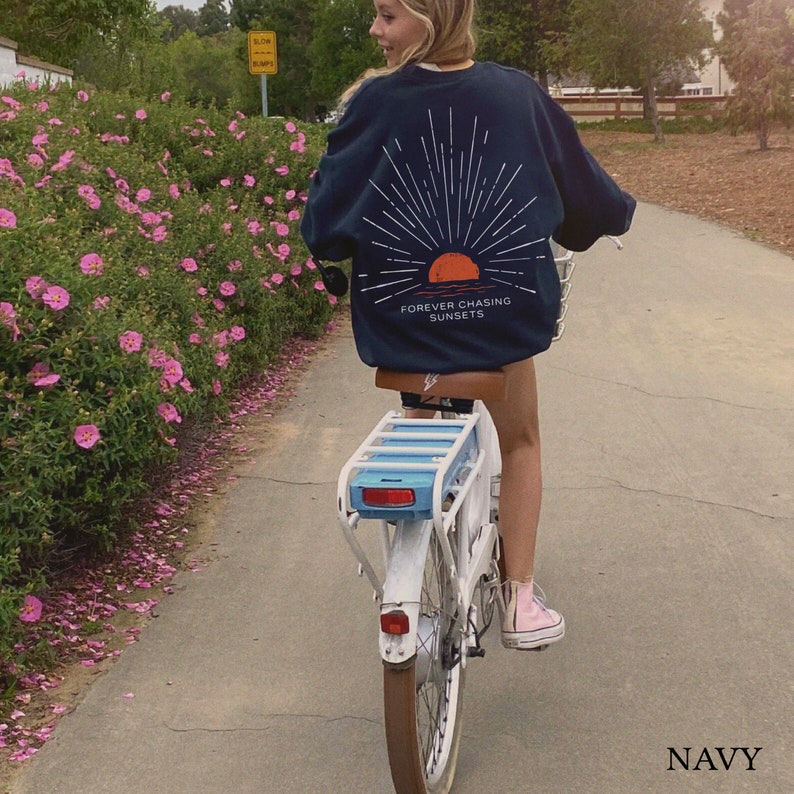 May include: A person riding a white electric bicycle with a blue basket. The person is wearing a navy blue sweatshirt with a graphic of a sunset and the text "FOREVER CHASING SUNSETS". The sweatshirt is navy blue.