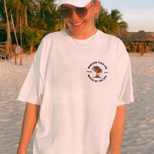 Puede incluir: Camiseta blanca con un estampado gr&aacute;fico de una palmera y el texto "Mightier than the waves of the sea".