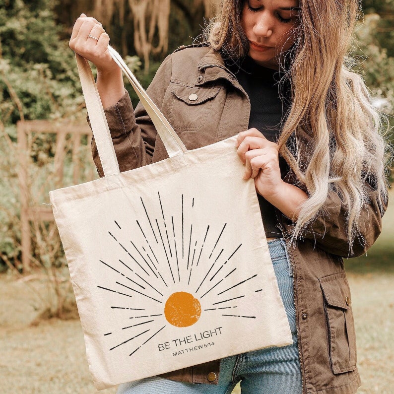 Op de afbeelding: Beige canvas draagtas met een zonnestralenontwerp. Een oranje cirkel en zwarte lijnen vormen de zon, met de tekst "BE THE LIGHT" en "MATTHEW 5:14" eronder. De tas heeft lange, beige handvatten.