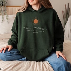 Pu&ograve; includere: Felpa con cappuccio verde bosco con una grafica di un sole che tramonta sulle onde e il testo "Through Deep Waters I Will Be With You, Joshua 1:9".