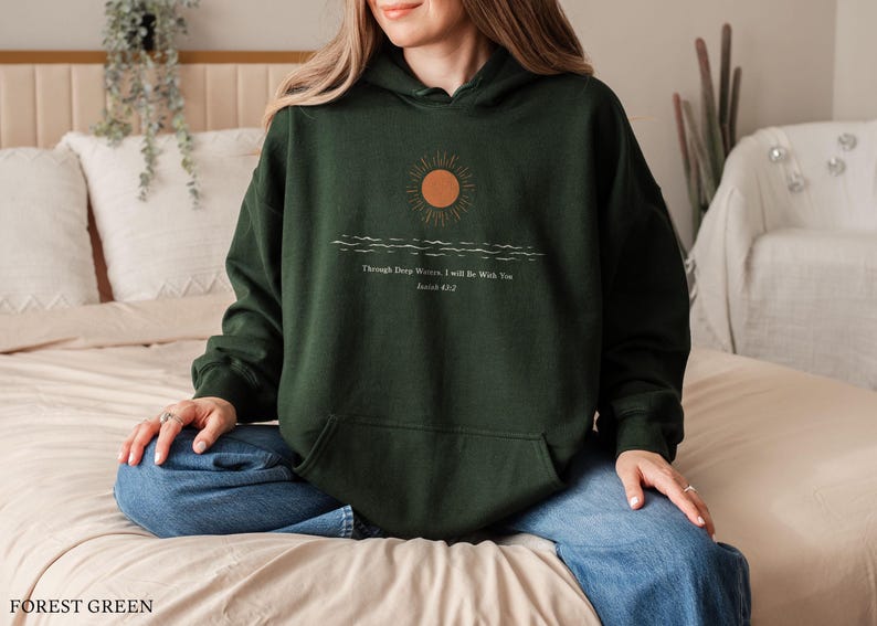 Pu&ograve; includere: Felpa con cappuccio verde bosco con una grafica di un sole che tramonta sulle onde e il testo "Through Deep Waters I Will Be With You, Joshua 1:9".