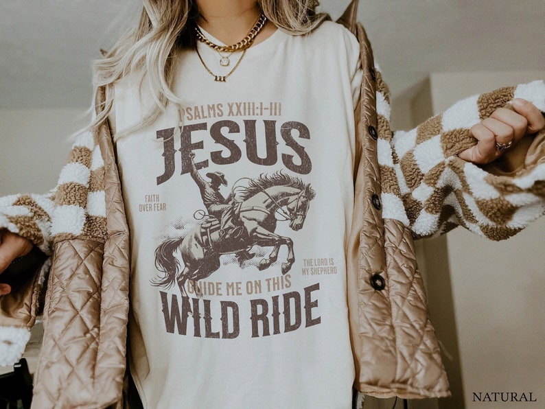 K&ouml;nnte beinhalten: Ein cremefarbenes T-Shirt mit einer braunen Grafik eines Cowboys, der ein Pferd reitet. Die Grafik enth&auml;lt den Text "PSALMS XXIII:I-III JESUS", "FAITH OVER FEAR", "GUIDE ME ON THIS WILD RIDE" und "THE LORD IS MY SHEPHERD".