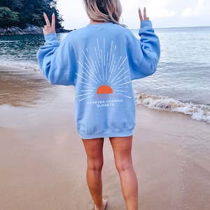 May include: A light blue sweatshirt with a graphic of a sunset and the text "FOREVER CHASING SUNSETS". The sweatshirt is worn by a person standing on a sandy beach.