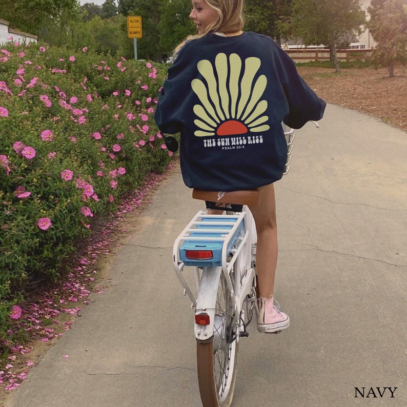 May include: A person riding a blue and white bicycle with a navy blue sweatshirt that says "The Sun Will Rise, Psalm 30:5". The sweatshirt has a yellow sun graphic with a red center.