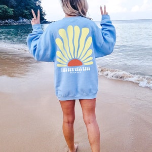 May include: A light blue sweatshirt with a yellow sun graphic and the text "The Sun Will Rise, Psalm 30:5" on the back. The sweatshirt is being worn by a person standing on a sandy beach.