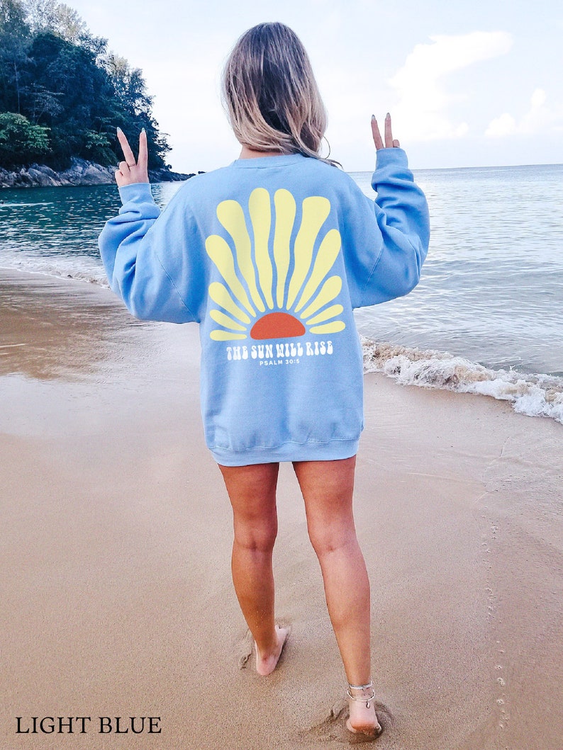 May include: A light blue sweatshirt with a yellow sun graphic and the text "The Sun Will Rise, Psalm 30:5" on the back. The sweatshirt is being worn by a person standing on a sandy beach.