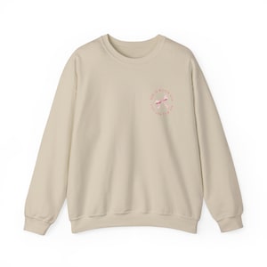 May include: A beige sweatshirt with a pink and white bow graphic on the front. The graphic is a circle with the text "God is within reach" written around the bow.