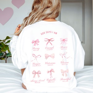 May include: White sweatshirt with the text "GOD SAYS I AM" printed on the back. The text is surrounded by pink bows with bible verses printed below each bow. The verses are: Strong (Phil 4:13), Enough (2 Cor 12:9), Brave (Deut 31:6), Unique (Psalm 139:13), Victorious (Rom 8:37), Chosen (Deut 31:6), Always Loved (Rom 8:38), Capable (Mark 10:27), Beautiful (Ecc 3:11).