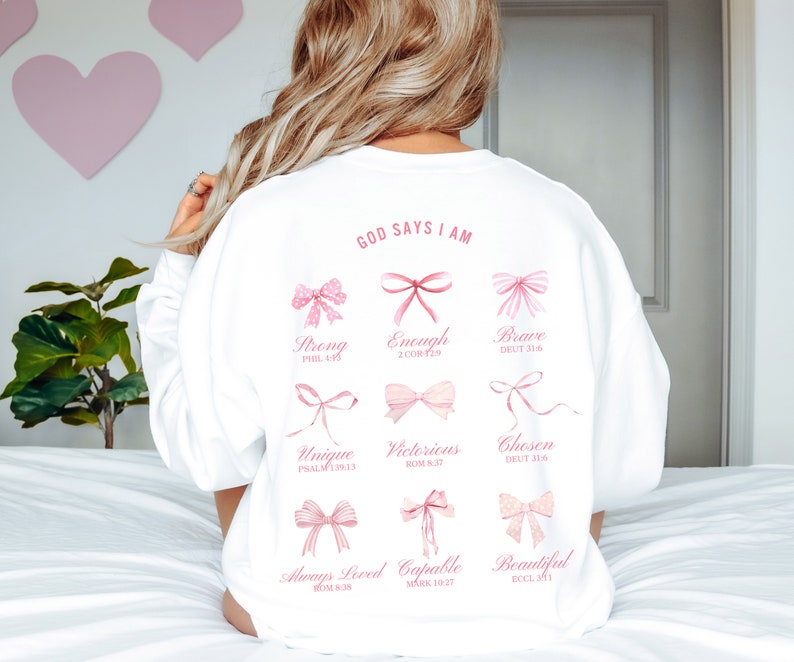 May include: White sweatshirt with the text "GOD SAYS I AM" printed on the back. The text is surrounded by pink bows with bible verses printed below each bow. The verses are: Strong (Phil 4:13), Enough (2 Cor 12:9), Brave (Deut 31:6), Unique (Psalm 139:13), Victorious (Rom 8:37), Chosen (Deut 31:6), Always Loved (Rom 8:38), Capable (Mark 10:27), Beautiful (Ecc 3:11).