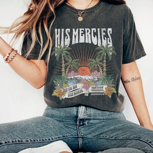 May include: A dark gray t-shirt with a vintage graphic design featuring palm trees, a sunset, and the text "His Mercies". The graphic also includes the text "They are new each morning" and "Great is your faithfulness".