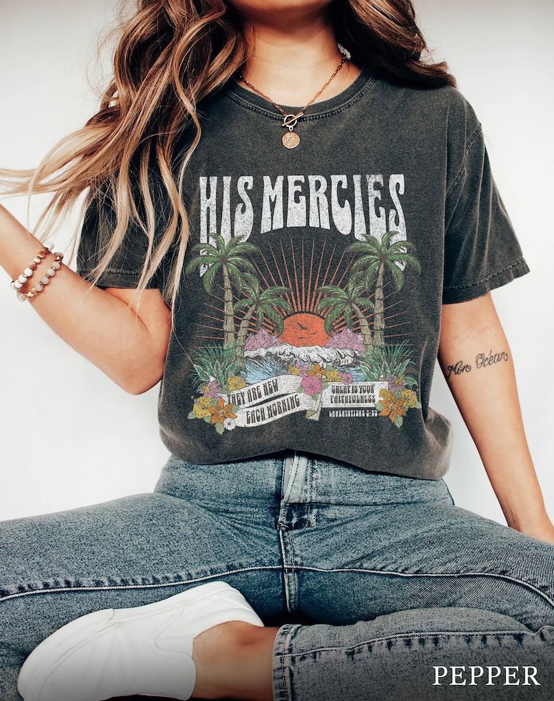 May include: A dark gray t-shirt with a vintage graphic design featuring palm trees, a sunset, and the text "His Mercies". The graphic also includes the text "They are new each morning" and "Great is your faithfulness".
