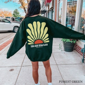 May include: A black sweatshirt with a yellow and orange sun graphic and the text "The Sun Will Rise, Psalm 30:5". The sweatshirt is shown on a model in front of a brick building.