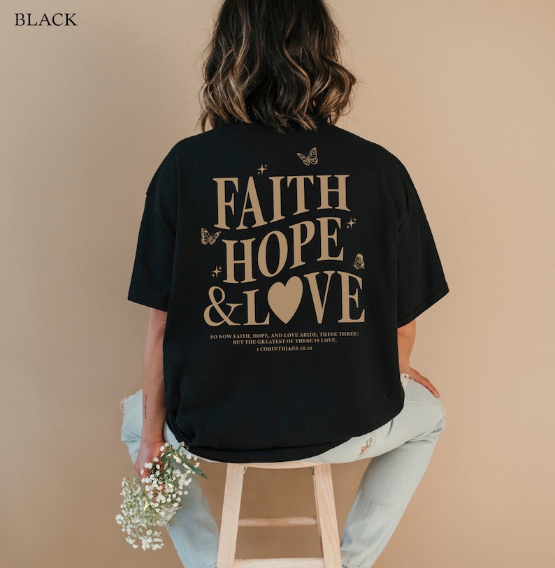 May include: Black t-shirt with a graphic print of the words "Faith Hope & Love" in a beige color with a heart and butterflies. The text "So now faith, hope, and love abide, these three; but the greatest of these is love. 1 Corinthians 13:13" is printed below the graphic.