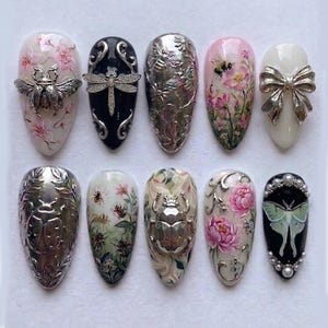 May include: A collection of ten intricately designed artificial nails. Each nail features unique artwork, including floral patterns, insects, and metallic embellishments. Colors include pink, white, black, and silver, showcasing a variety of artistic styles and textures.