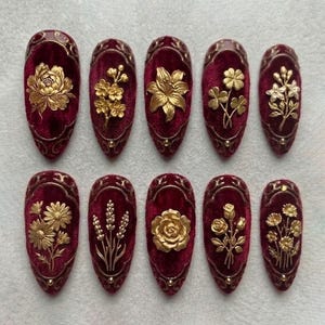 Könnte beinhalten: Ein Set aus zehn oval geformten, weinroten Kunstnägeln. Jeder Nagel zeigt ein goldenes Blumendesign, darunter Rosen, Lilien und andere detaillierte botanische Elemente. Die Nägel haben eine samtartige Textur und einen dekorativen Goldrand.