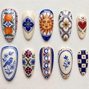 May include: A collection of ten almond-shaped nail art designs. Each nail features a unique hand-painted design, including oranges, a sun face, floral patterns, and a blue bird. The color palette includes white, blue, red, and gold.