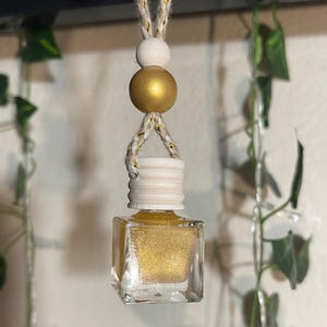May include: A hanging car air freshener with a square glass bottle filled with gold glitter. The bottle is topped with a white cap and a wooden bead. The air freshener is suspended by a beige rope with gold accents.